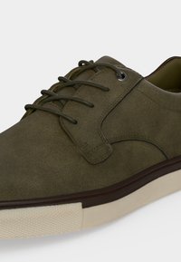 Chaussure à lacets en daim vert olive avec une bordure marron foncé et une semelle en caoutchouc ivoire, conçue pour un usage décontracté.