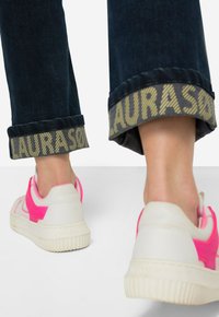 Sneakers bianchi con dettagli rosa e suola in gomma, abbinati a jeans blu scuro con un risvolto a fantasia contrastante.