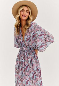 Maxi šaty s kvetinovým vzorom v svetlom modrom odtieni a ružovým paisley dizajnom; majú hlboký výstrih do V a elastický pás. Model má na hlave široký klobúk.