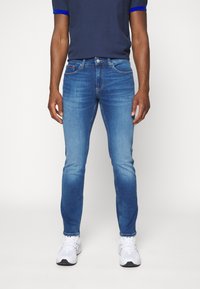 Modré džíny ze džínoviny s úzkým střihem, obsahující vybledlé části, pět kapes a klasické zapínání na knoflíky, v kombinaci s bílými sportovními botami.