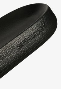 Zwarte rubberen sandaal met een gestructureerd oppervlak, voorzien van een reliëf "SUPERDRY" logo op de zijkant, dat een minimalistisch en sleek ontwerp laat zien.