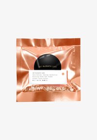 Kupfertasche mit transparenter Vorderseite, die unscharfes Puder für die Augenpartie zeigt. Schwarzes rundes Etikett mit goldener Schrift und weißer Produktbeschreibung.