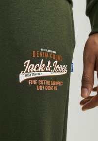 Sötétzöld pamut ruházat, fehér és narancssárga hímezett felirattal, amely tartalmazza a "Jack & Jones" és a "Denim Goods" kifejezéseket. Simára váltott textura.