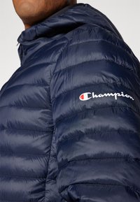 Jūras spēku pūkains jaka, izgatavota no spīdīgas auduma. Dizaņā ir kapuce, kviltots dizains un balta "Champion" logo kreisajā plecā.