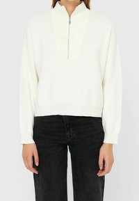 Witte ritsvest met hoge kraag en gestructureerde breiwerk, cropped op de taille, gecombineerd met zwarte high-waisted jeans.