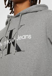 Szary hoodie z sznurkiem, z wyraźnym czarnym logo "CK" oraz białym napisem "Calvin Klein Jeans" z przodu, wykonany z miękkiego materiału.