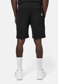 Pantalones cortos de sudor negro con cintura elástica, bolsillos laterales y un pequeño logotipo rojo y blanco en el lado izquierdo. Llevados con calcetas y zapatos blancos.