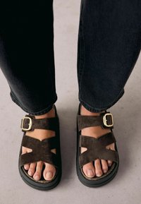 Sandale din suede maro cu design încrucișat, detaliu cu cataramă aurie, talpă neagră ridicată și degete expuse. Purtați cu blugi scurți închis.
