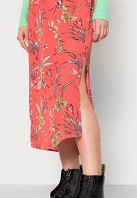 Robe florale corail avec une fente latérale, ornée de motifs de fleurs vibrants. Associée à des bottines noires vernies. Finition en tissu texturé.