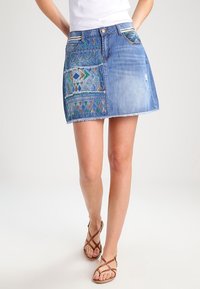 Mini-jupe en jean avec broderie géométrique colorée sur un côté, ourlet effiloché et conception traditionnelle à cinq poches.