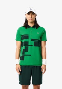Grünes Polohemd mit schwarzen geometrischen Mustern, kurzen Ärmeln und einem schwarzen Kragen. Kombiniert mit dunkelgrünen Shorts und einer weißen Kappe.