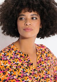 Mehrfarbige Bluse mit einem lebhaften abstrakten Muster in Orange, Pink und Schwarz. Weicher Stoff mit V-Ausschnitt und Knopfdetailed.
