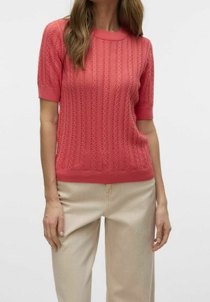 Vrouw met een koraalroze tricot shirt met korte mouwen en verticale patronen, en beige broek met hoge taille, staand tegen een effen achtergrond.