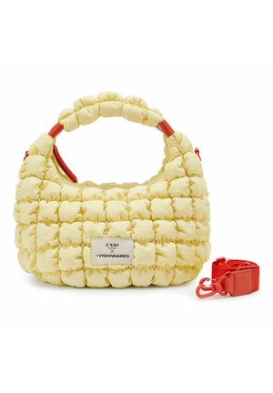 Pequeño bolso acolchado amarillo con asa acolchada, etiqueta frontal y correa roja para el hombro desmontable colocada junto a él.