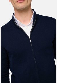 Homme portant un pull zippé bleu marine sur une chemise blanche, vu de l'épaule au menton sur un fond blanc.