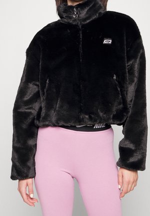 Zwarte zachte jas met rits en hoge kraag gedragen over een zichtbare zwarte Nike tailleband, gecombineerd met lichtroze strakke leggings.