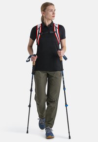 Maglietta polo nera, pantaloni verde oliva, bastoni da escursionismo, scarpe blu. Cinghie rosse e bianche sullo zaino. Postura eretta, espressione concentrata.