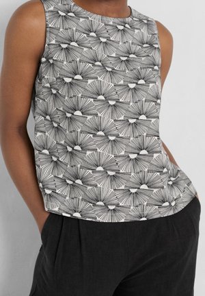 Sleeveless black and white top with geometric sunburst pattern worn with black pants, hands in pockets, against plain background.