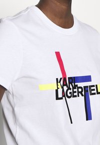 Bílý bavlněný tričko s grafickým designem v sytých barvách: červená, modrá, žlutá a černý text "KARL LAGERFELD". Krátké rukávy.
