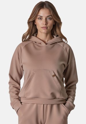Jonge vrouw met lichtbruin haar die een beige capuchontrui en bijpassende broek draagt, staand met handen in de zakken.