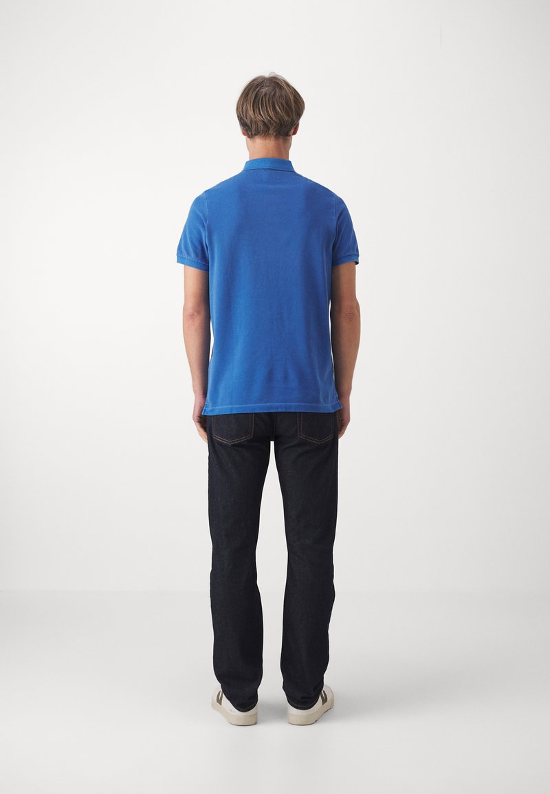 Camisa polo de algodón azul con mangas cortas, corte recto, combinada con jeans de mezclilla oscuros y zapatillas blancas, vista desde la espalda.