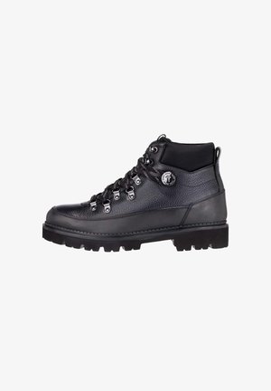Bota de senderismo de cuero negro con ojetes metálicos, suela gruesa con dibujo, cuello acolchado y placa redonda con logo en el tobillo.