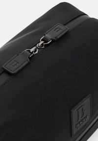 Svart nylonväska med en elegant design, accentuerad med en metallblixtlås och ett strukturerat logopatch. Slät yta med minimalistiska detaljer.