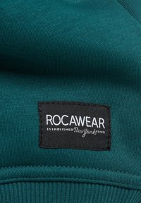 Türkiz kapucnis pulóver anyag bordázott textúrával. Fekete címkével rendelkezik, amelyen fehér betűkkel olvasható: "ROCAWEAR ESTABLISHED NewYork 1999."