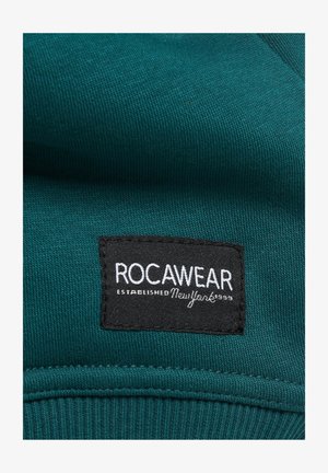 Türkiz kapucnis pulóver anyag bordázott textúrával. Fekete címkével rendelkezik, amelyen fehér betűkkel olvasható: "ROCAWEAR ESTABLISHED NewYork 1999."