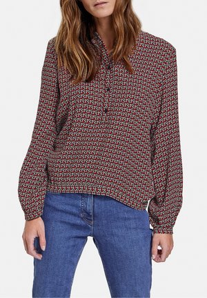 Femme portant un chemisier à manches longues à motifs rouges, blancs et noirs, associé à un jean bleu sur fond blanc.
