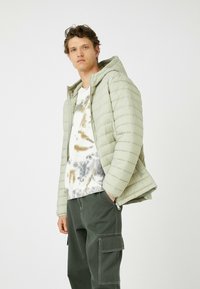 Lys grøn pufferjakke med hætte, vandret quiltning, parret med en hvid og grå tie-dye t-shirt og mørke cargo-bukser med lommer.