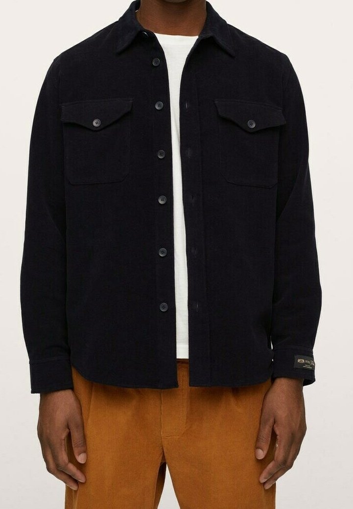 Homme portant une veste-chemise noire à boutons sur un t-shirt blanc et un pantalon en velours côtelé marron, debout, les bras détendus le long du corps.
