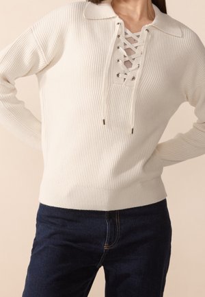 Femme portant un pull en maille côtelée crème avec un laçage devant et un col, associé à un jean bleu foncé.