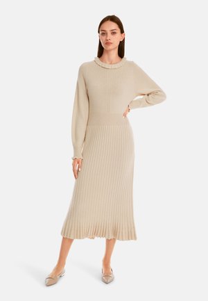 Langes, geripptes Strickkleid in Beige, mit Rüschenausschnitt und tailliertem Oberteil sowie ausgestelltem Saumabschluss. Das Modell trägt spitze Ballerinas.