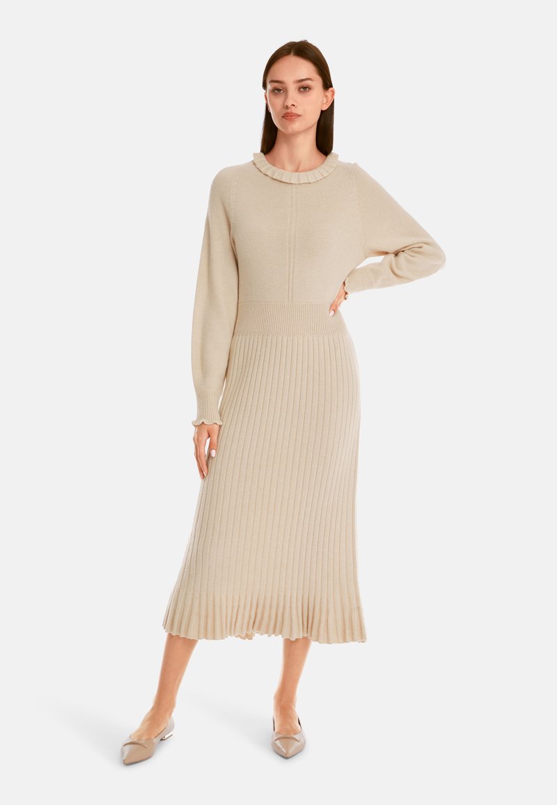 Langes, geripptes Strickkleid in Beige, mit Rüschenausschnitt und tailliertem Oberteil sowie ausgestelltem Saumabschluss. Das Modell trägt spitze Ballerinas.