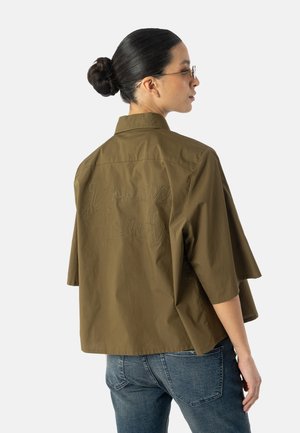 Kvinde med håret sat op i en knold, iført briller, en oversized olivengrøn skjorte med teksten "Dream loud" broderet på ryggen og blå jeans, set fra siden.