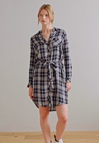 Abito a camicia a quadri in blu navy e bianco, con una chiusura frontale a bottoni, vita con laccio, maniche lunghe e una vestibilità morbida. Tessuto dalla texture liscia.