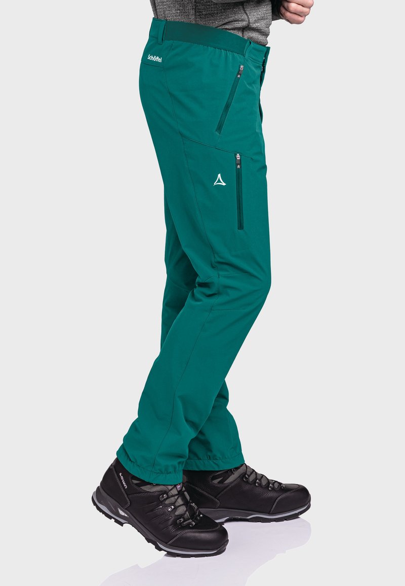 Teal-Funktionshose aus Softshell mit Reißverschlusstaschen, elastischem Bund und verstärkten Nähten, kombiniert mit schwarzen Wanderschuhen mit Schnürsenkeln und strukturierten Sohlen.