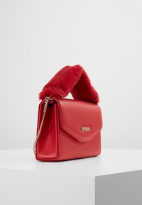 Červená kožená kabelka s plyšovým červeným umělým kožíškem na uchu, řetízkovým popruhem a zlatou logovou destičkou na přední straně. Ostrý design, strukturovaný tvar.