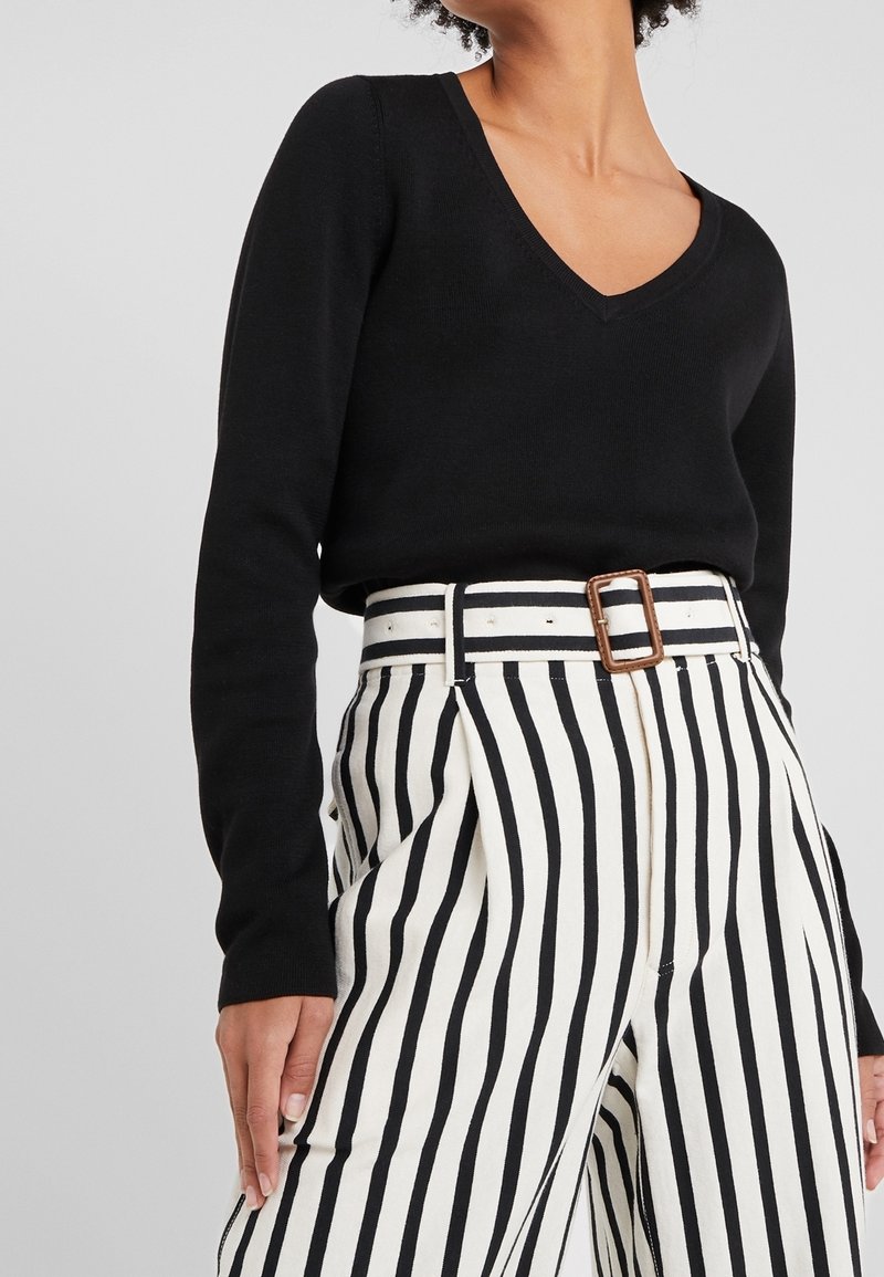 Pull noir à manches longues avec un décolleté en V, associé à un pantalon taille haute blanc avec des rayures verticales noires et une boucle de ceinture rectangulaire.