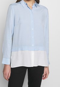 Camicia a righe blu chiaro e bianca con maniche lunghe, apertura frontale con bottoni, colletto e orlo inferiore bianco, realizzata in un tessuto morbido e leggero.
