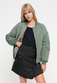 Veste bomber verte avec fermeture éclair orange, associée à une jupe matelassée noire avec détails de boutons et poche latérale. Haut noir uni en dessous.