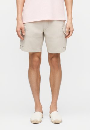 Beige cargoshorts med klaplommer båret af en stående person i hvide slip-on sko og en lyserød skjorte, mod en ensfarvet baggrund.
