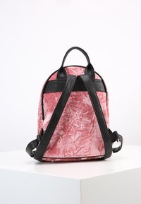 Mochila de terciopelo rosa con correas y adornos de cuero negro. Forma redondeada, superficie texturizada y doble asa en la parte superior para llevar.