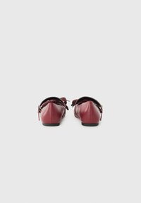 Burgundy nahkaharsokkeet pyöreällä varvaskohdalla, mustalla pohjalla ja koristeellisen nauhan yksityiskohdalla takana. Suuri tekstuuri, minimalistinen muotoilu.