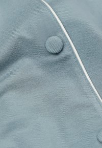 Světle modrá látka s ribovanou texturou, výrazný kulatý knoflík a kontrastní bílá lemovina podél okraje.