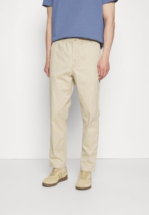 Man draagt beige broek met rechte pijpen, beige sneakers en een blauw shirt met korte mouwen tegen een effen witte achtergrond.