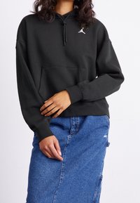 Sudadera negra con capucha, bolsillo delantero y cordón, con un logo blanco. Combinada con una falda vaquera azul que muestra una tela con textura.