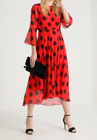 Robe rouge à pois blancs avec manches transparentes, taille froncée et longueur midi. Accessoirisée avec une pochette noire et des talons noirs à brides.