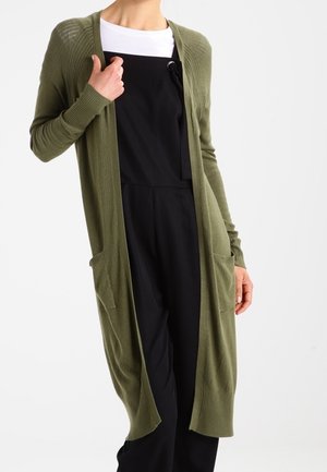 Person iført en lang olivengrøn cardigan med åben front over en hvid top og sorte bukselignende tøj, med hænderne tæt på lommerne og brystet.